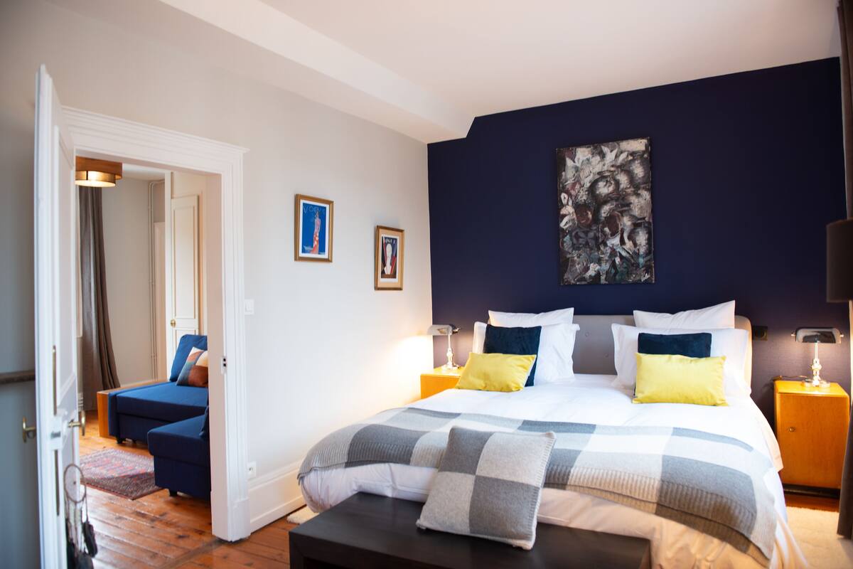 A bedroom with a large bed, a bedside table, a nightstand, a blue sofa, and a door leading to another room.