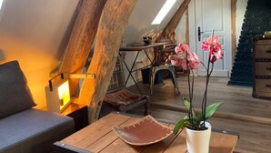 Un dormitorio estilo loft acogedor con vigas de madera a la vista, una mesa de madera con posavasos de cuero, una planta en maceta y una cama con cabecero gris.