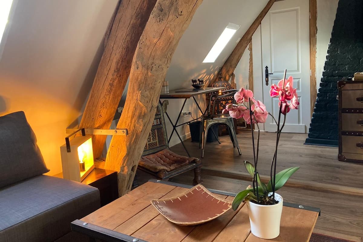 A cozy loft bedroom with exposed wooden beams, a wooden table with a leather placemat, a potted plant, and a bed with a grey headboard.
