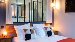 Una cama bien hecha con sábanas blancas y una manta roja, una mesita de noche de madera con una lámpara, y un espejo grande sobre la cama.