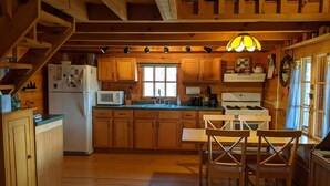 A wooden cabin kitchen with a dining table and chairs, a refrigerator, microwave, stove, and a window with curtains. - Amish-Built Cabin in the Driftless Area (Boscobel)