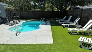 Un patio trasero con piscina, césped artificial y sillas de descanso blancas.