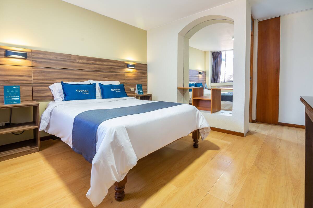 A hotel room with a wooden headboard, a bed with white and blue bedding, a nightstand, and a wooden floor.