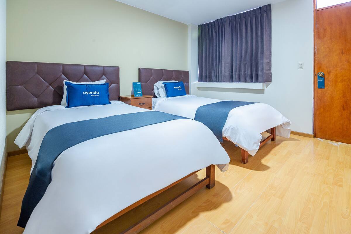 A hotel room with two beds, each with a headboard and a blue pillow with the hotel name. There is a wooden nightstand between the beds, and a window with curtains.