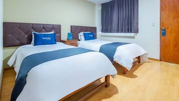 Habitación de hotel con dos camas, cada una con cabecera y almohada azul con el nombre del hotel. Hay una mesita de noche de madera entre las camas, y una ventana con cortinas.