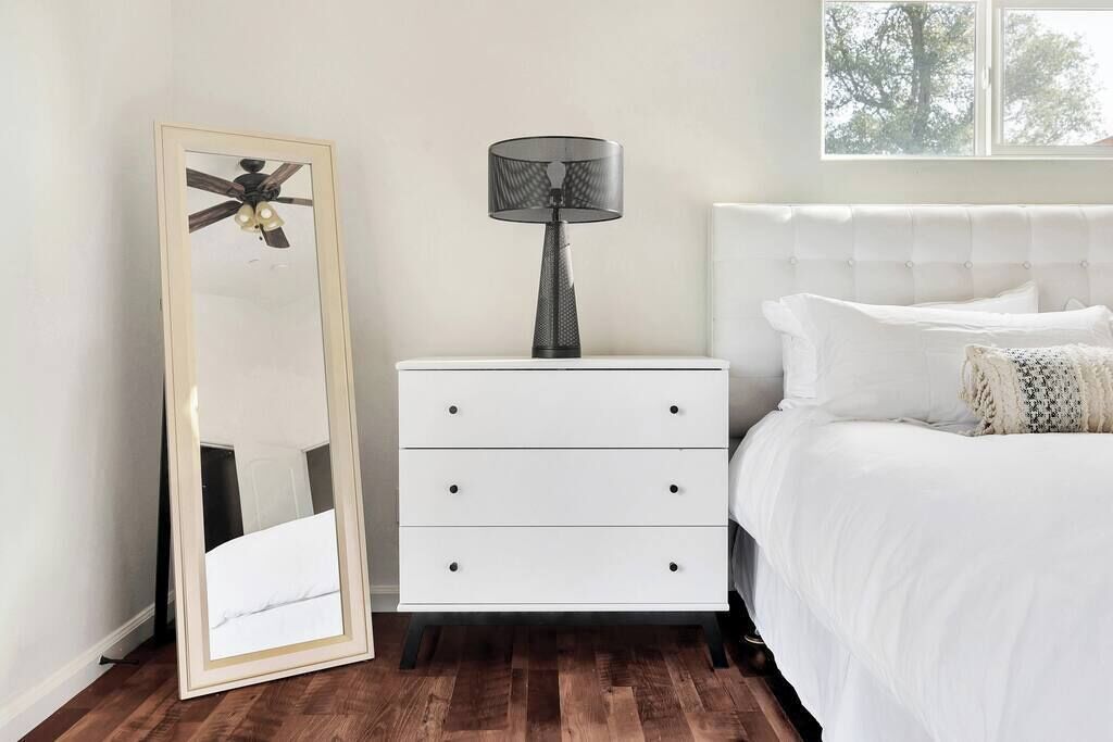 A bedroom with a white bed, a nightstand with a lamp, and a large mirror.