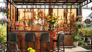 Un área de bar al aire libre con mostrador de madera, taburetes negros y un arreglo floral decorativo.