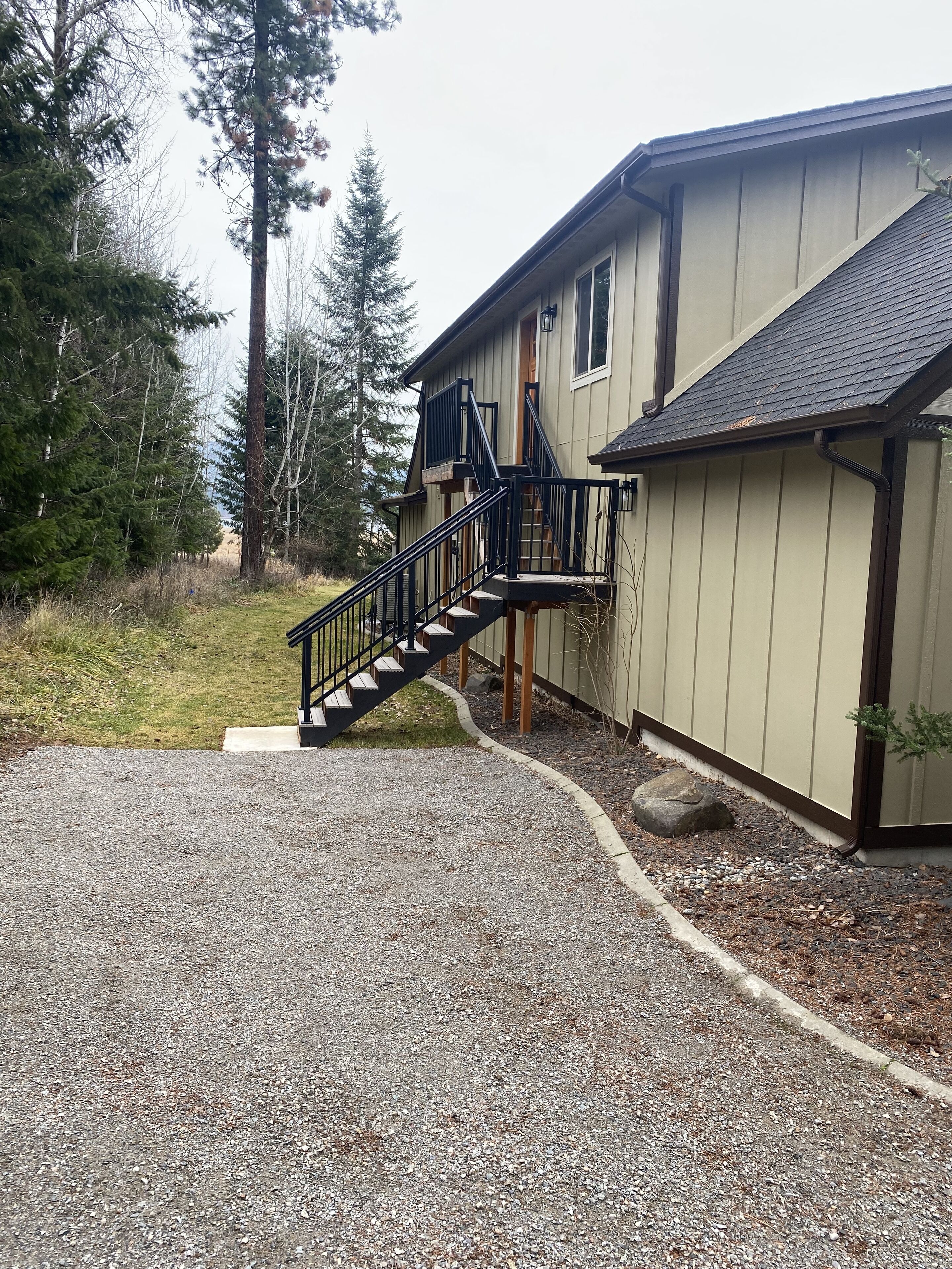 Une maison à deux étages avec une allée en gravier, une terrasse en bois et un escalier menant à la porte d’entrée.