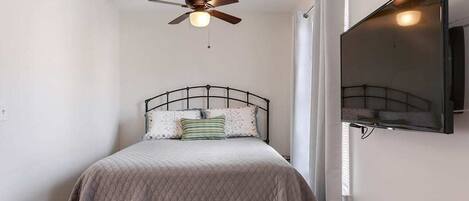 A bedroom with a bed, a ceiling fan, a mounted TV, and a window with curtains.