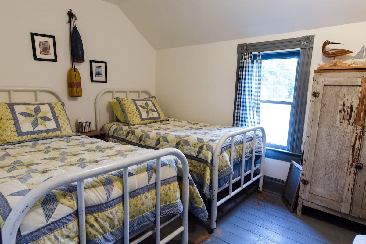 A room with two beds, each with a metal frame and quilted bedding. There are framed pictures on the wall, a window with a checkered curtain, and a wooden cabinet with a decorative item on top.