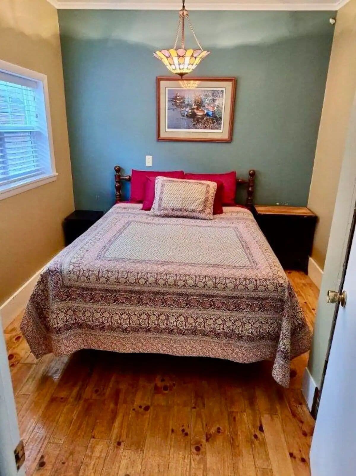 A bedroom with a bed, a chandelier, a framed picture on the wall, and a window with blinds.