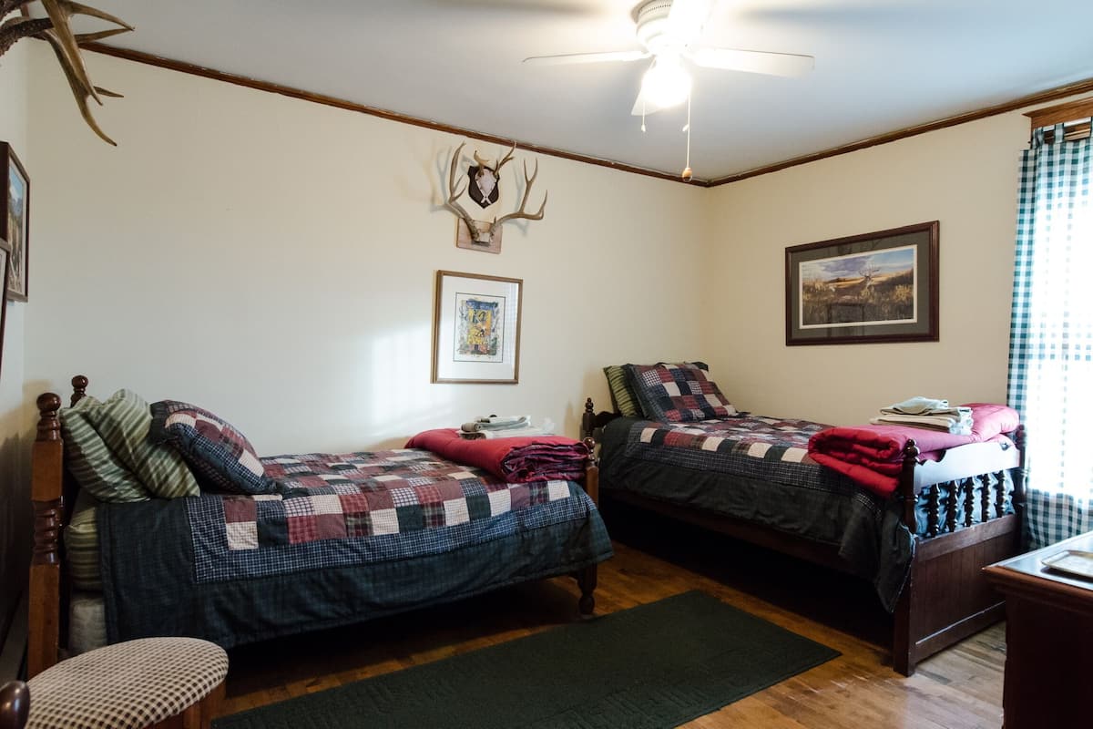 A room with two wooden beds, a ceiling fan, a framed deer head on the wall, and a framed landscape painting.