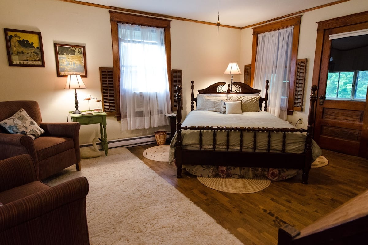A bedroom with a wooden bed, two bedside lamps, a green nightstand, a brown armchair, and a large window with curtains.