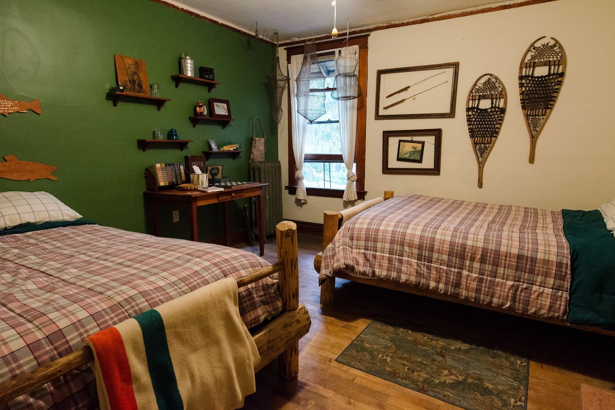 A room with two wooden beds, a small table, and a chair. There are shelves with various items and a window with curtains.