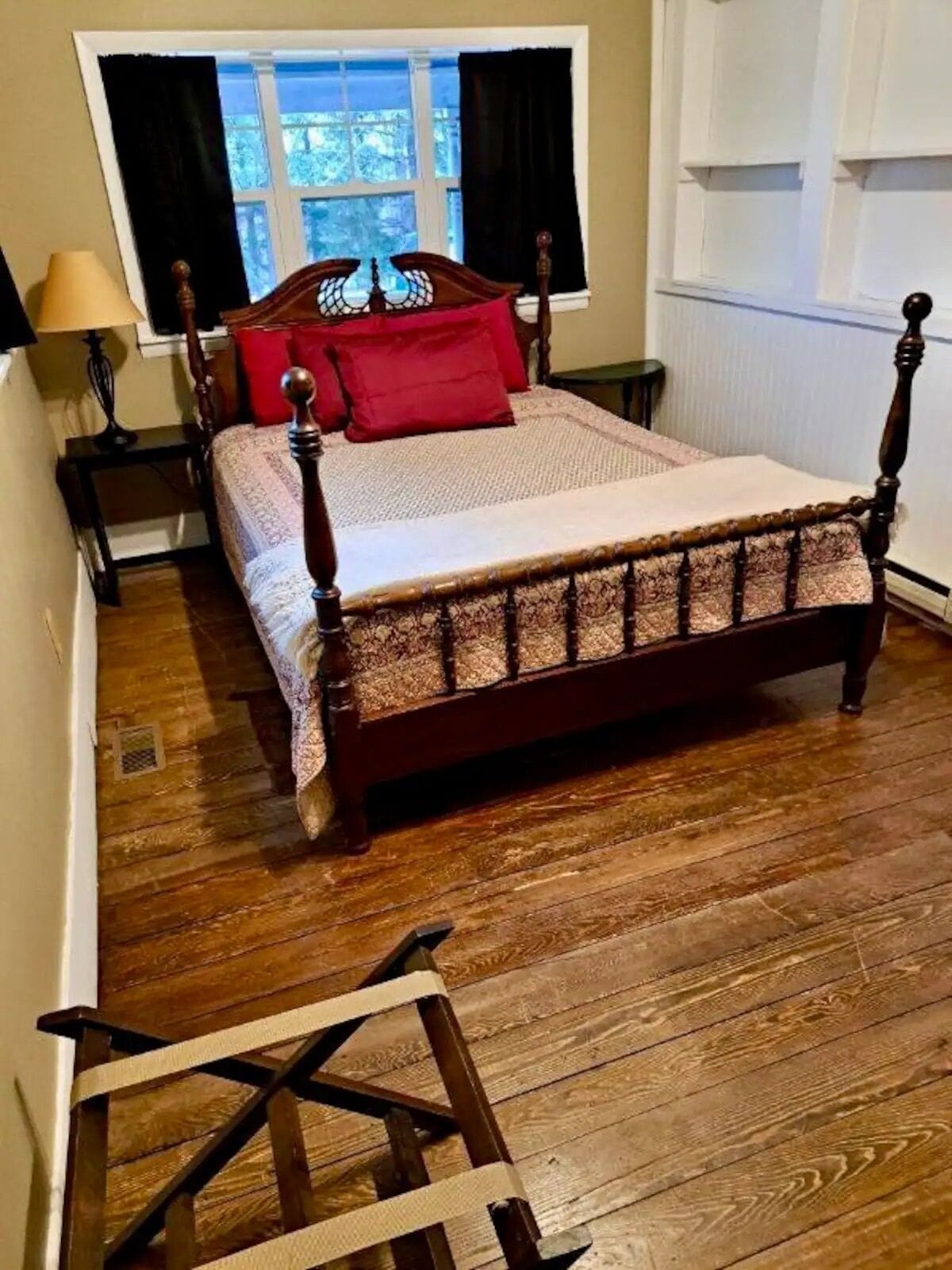 A bedroom with a wooden four-poster bed, a window with curtains, and a nightstand with a lamp.