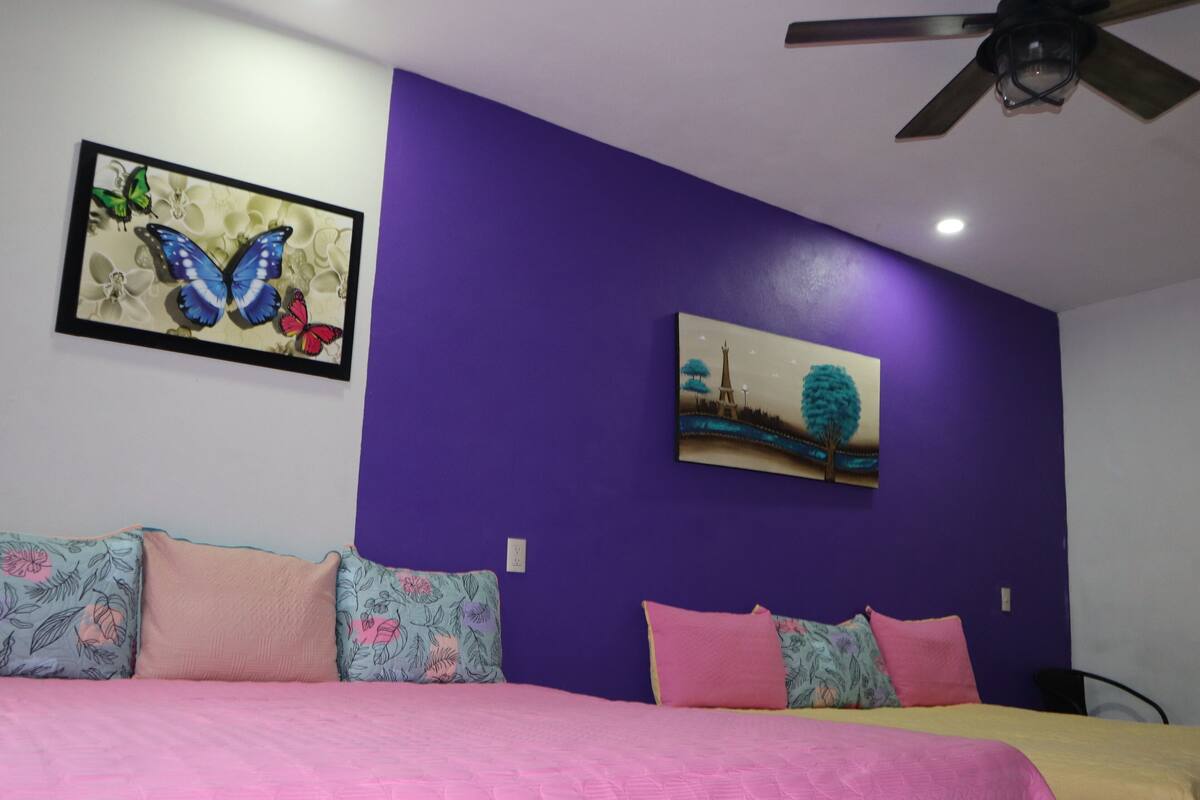 A bedroom with a purple wall, a bed with pink bedding, and two paintings.