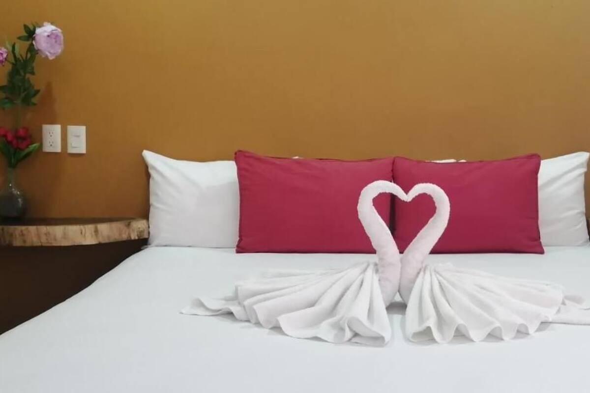 A hotel room with a bed, a heart-shaped towel arrangement, a vase with flowers, and a small sailboat decoration.