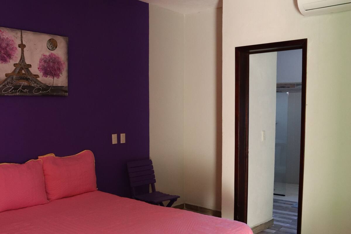 A bedroom with a large bed, purple walls, a ceiling fan, and a painting of the Eiffel Tower.