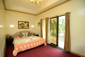 A bedroom with a bed, a painting, a nightstand, a window with curtains, and a view of greenery outside. - Sunny Point Hotel & Apartelle (Davao)
