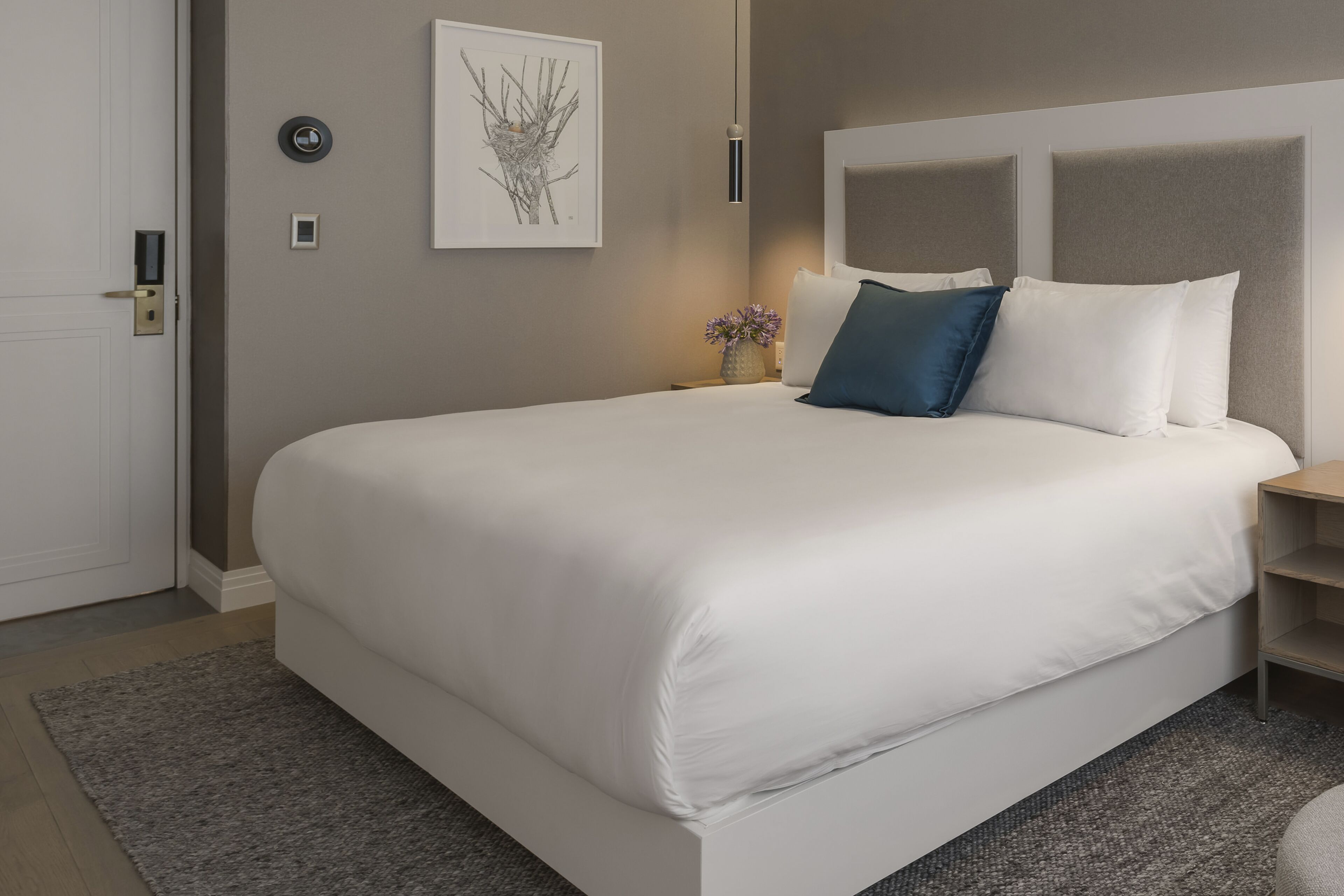 A modern hotel room with a large bed, a bedside table, a nightstand, a wall-mounted artwork, and a door with a lock.
