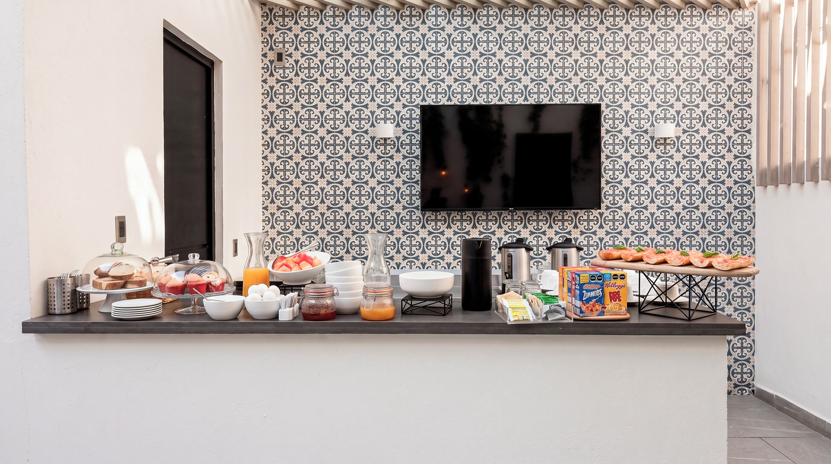 Uma mesa de pequeno-almoço com diversas opções de comida e uma televisão de ecrã plano na parede.