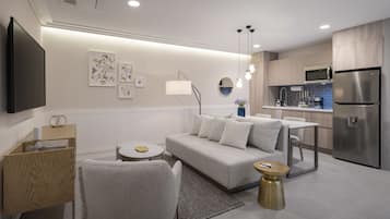 Una sala de estar moderna con sofá, mesa de centro y sillas, una cocina con refrigerador y microondas, y un televisor de pantalla plana montado en la pared.