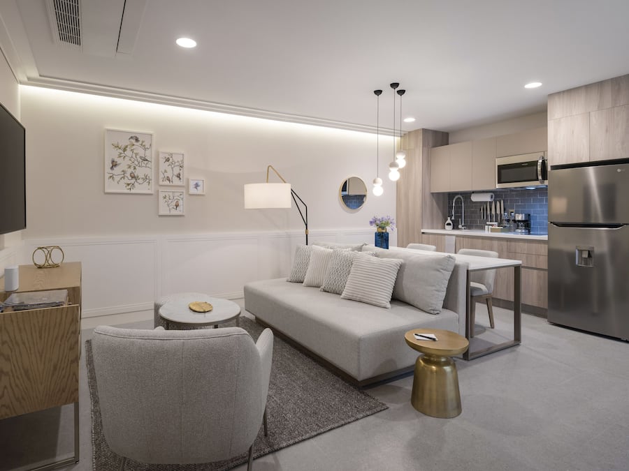 Una sala de estar moderna con sofá, mesa de centro y sillas, una cocina con refrigerador y microondas, y un televisor de pantalla plana montado en la pared.