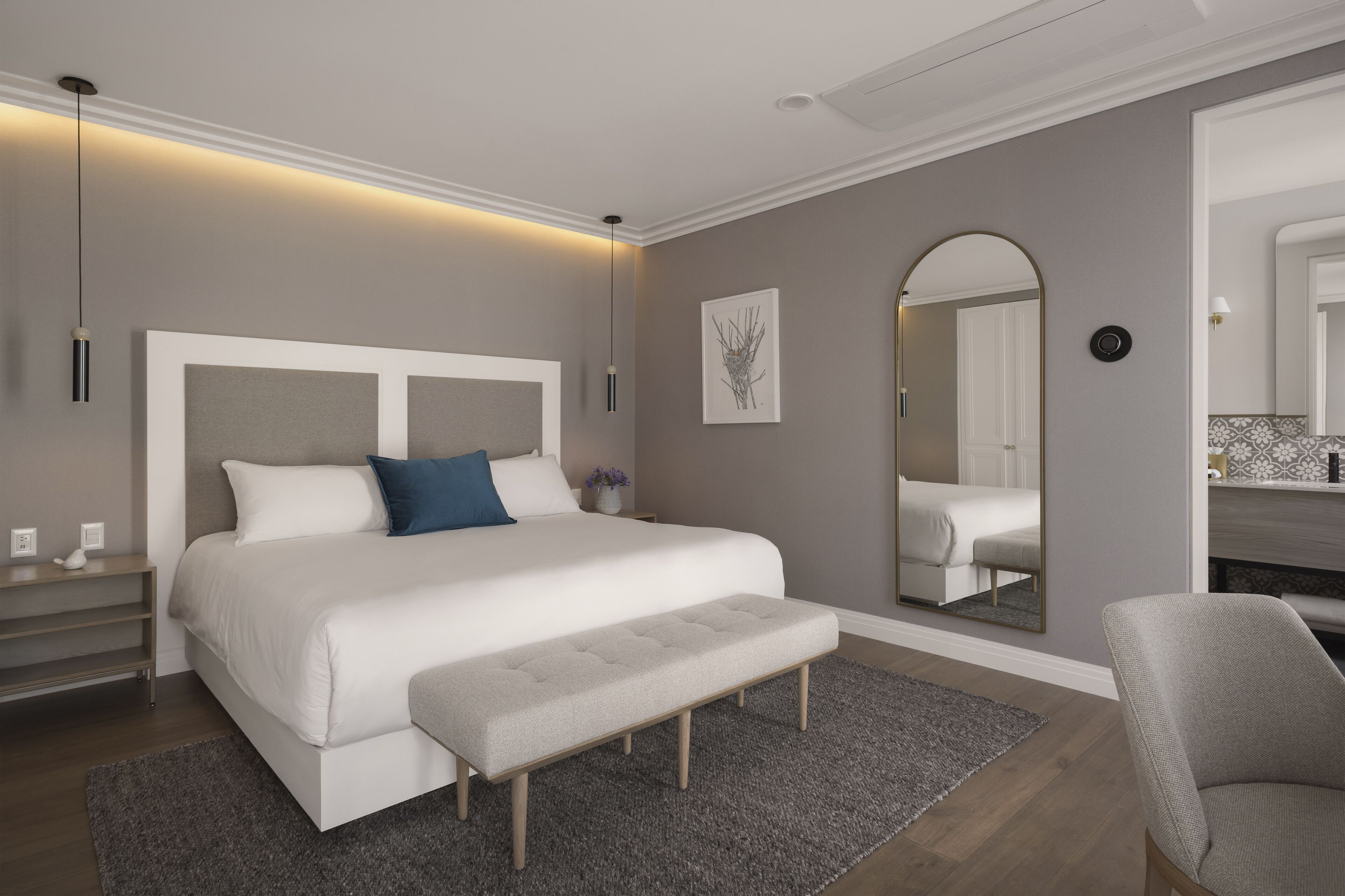 Um quarto moderno com uma cama grande, um banco, uma mesa de cabeceira, um espelho e um criado-mudo.