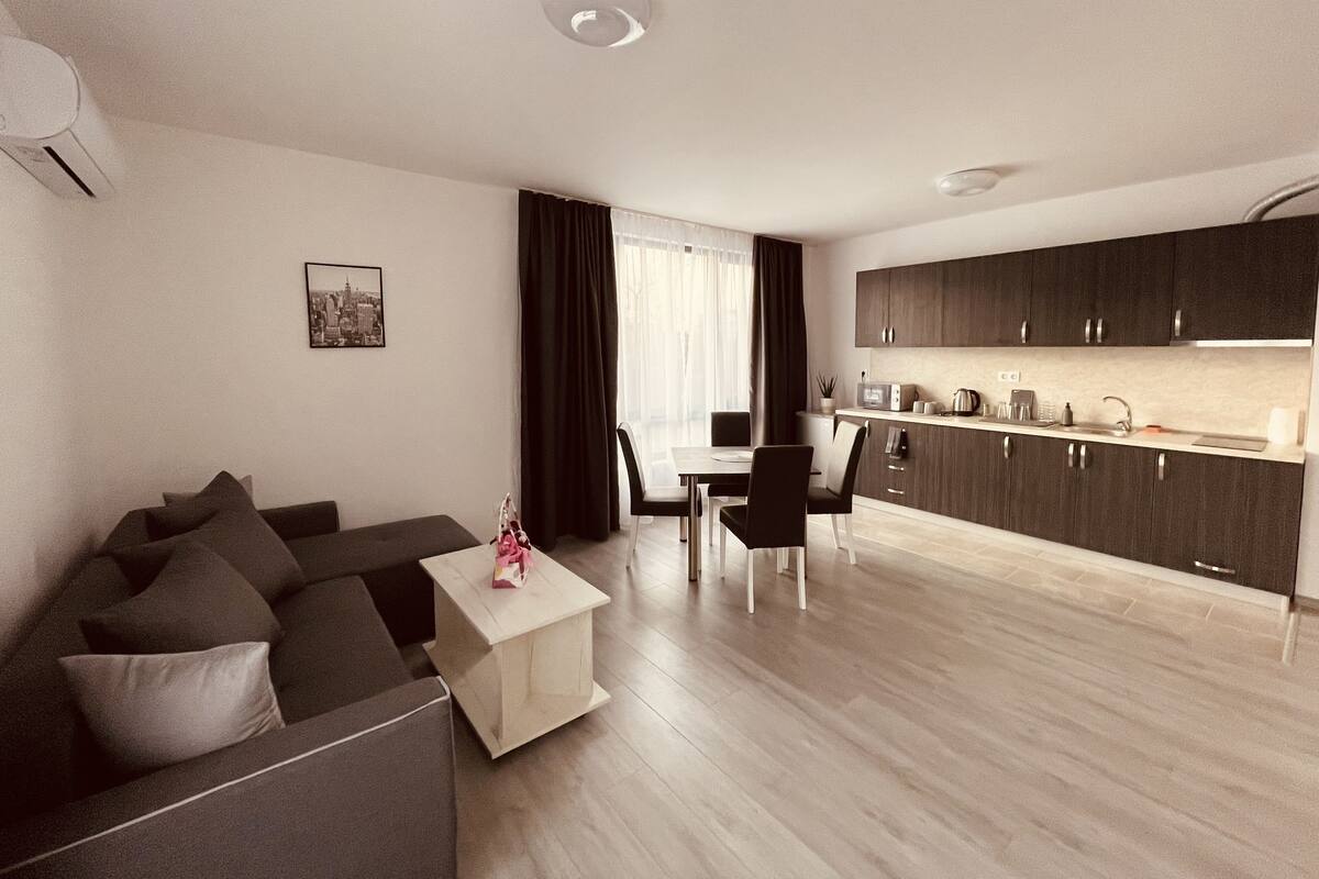 A modern living room with a sofa, a small coffee table, and a dining area with a table and chairs. A kitchen with dark cabinets and a countertop is visible in the background.