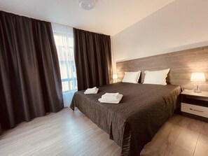 A hotel room with a large bed, a wooden headboard, a nightstand with a lamp, and a window with curtains. - ATM LUX APART (Sofia)