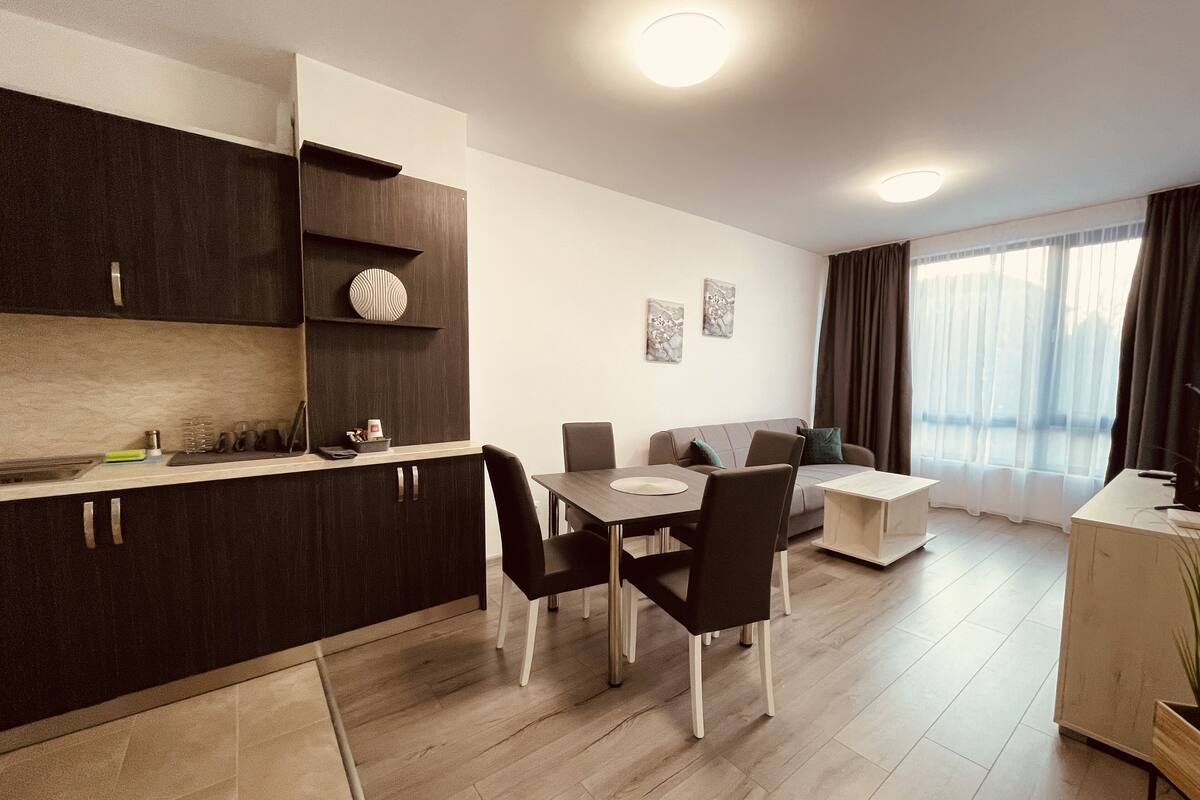 A modern kitchen with dark cabinetry, a dining area with a table and chairs, a sofa, and a large window with curtains.