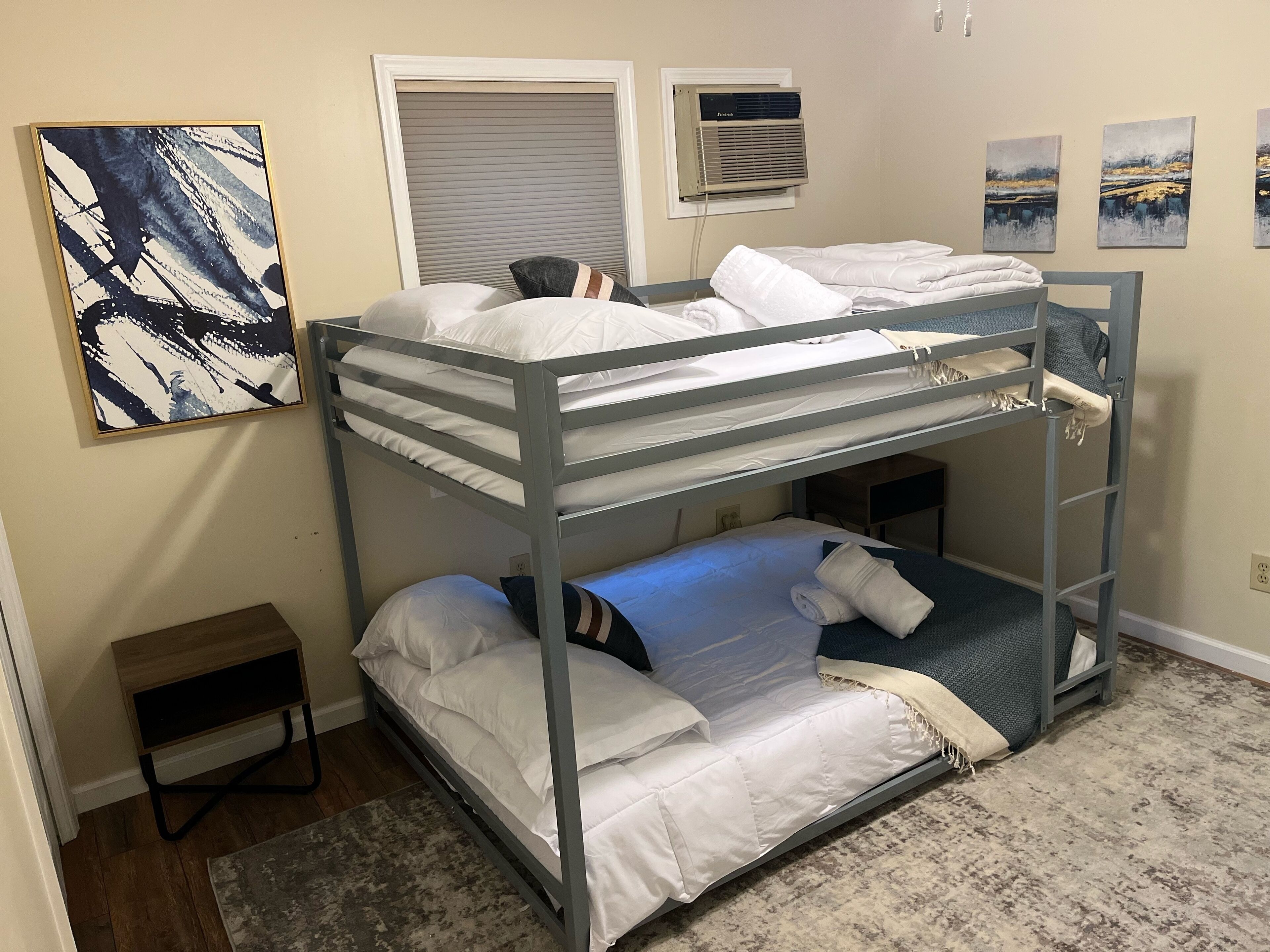 A bunk bed room with a small wooden side table, a window with blinds, and a wall-mounted air conditioner.