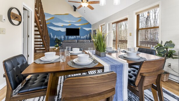 Un coin repas avec une table en bois dressée pour quatre personnes, entourée de chaises en cuir. Un ventilateur de plafond et des lampes suspendues se trouvent au-dessus de la table.