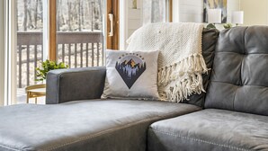 Un canapé en cuir gris avec un coussin blanc orné d’un motif représentant une montagne et une forêt, un plaid blanc à franges et une petite table ronde sur laquelle est posée une plante.