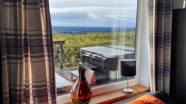 Una habitación con un ventanal grande que da a un paisaje verde, una terraza de madera y una lámpara negra en el alféizar.