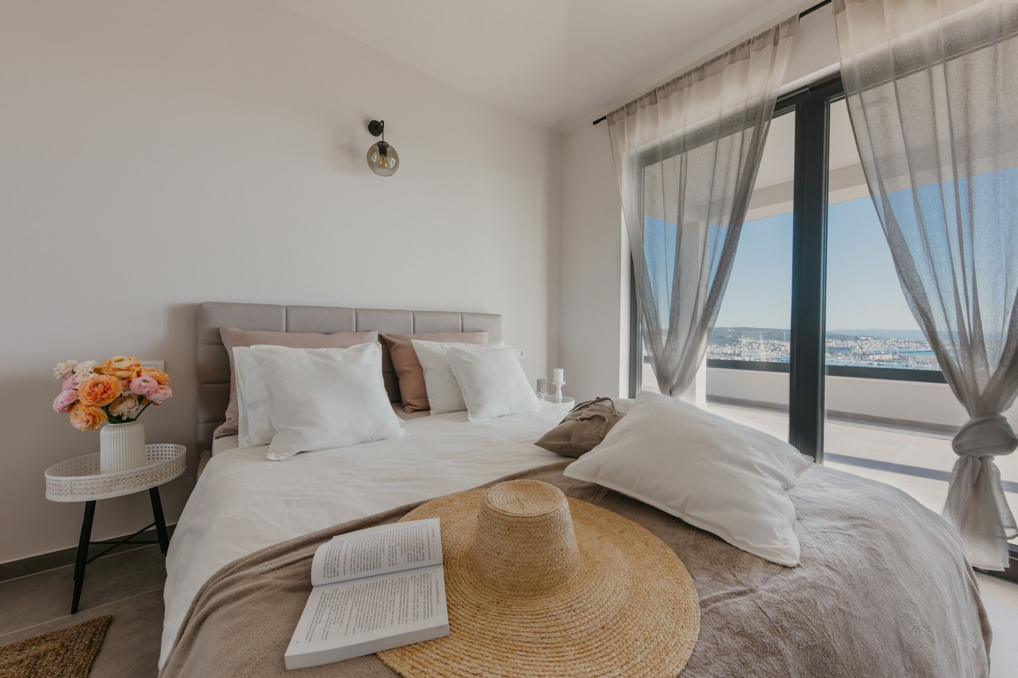 Un dormitorio con una cama grande, una mesita de noche con un jarrón de flores y vistas a la ciudad a través de cortinas traslúcidas.