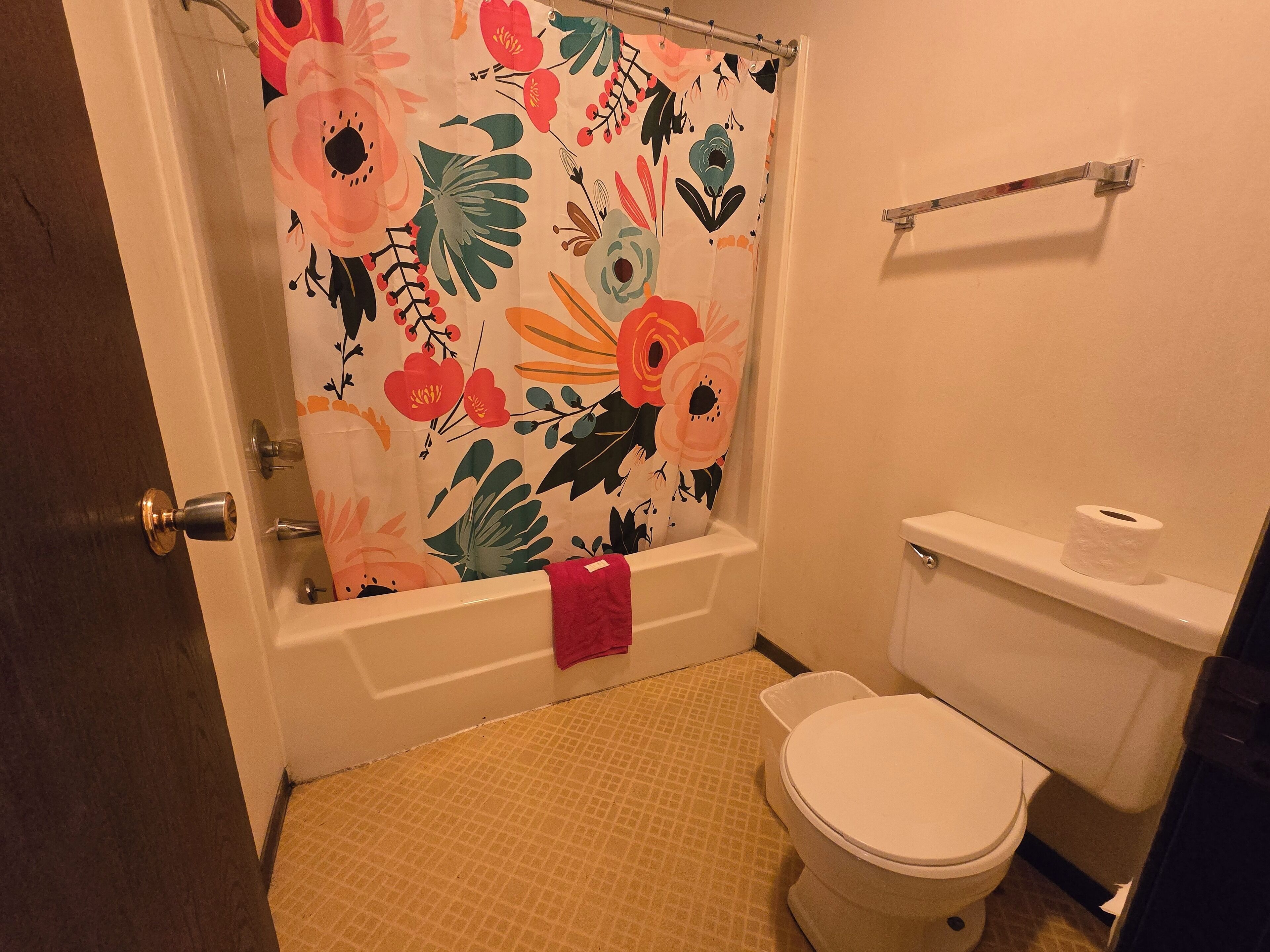 Un baño con cortina de ducha estampada con motivos florales, un inodoro blanco y una bañera con una toalla rosa.