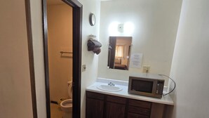 A small bathroom with a toilet, sink, and microwave. There is a mirror above the sink and a clock on the wall. - OYO Sandman Inn (Eagle Grove)