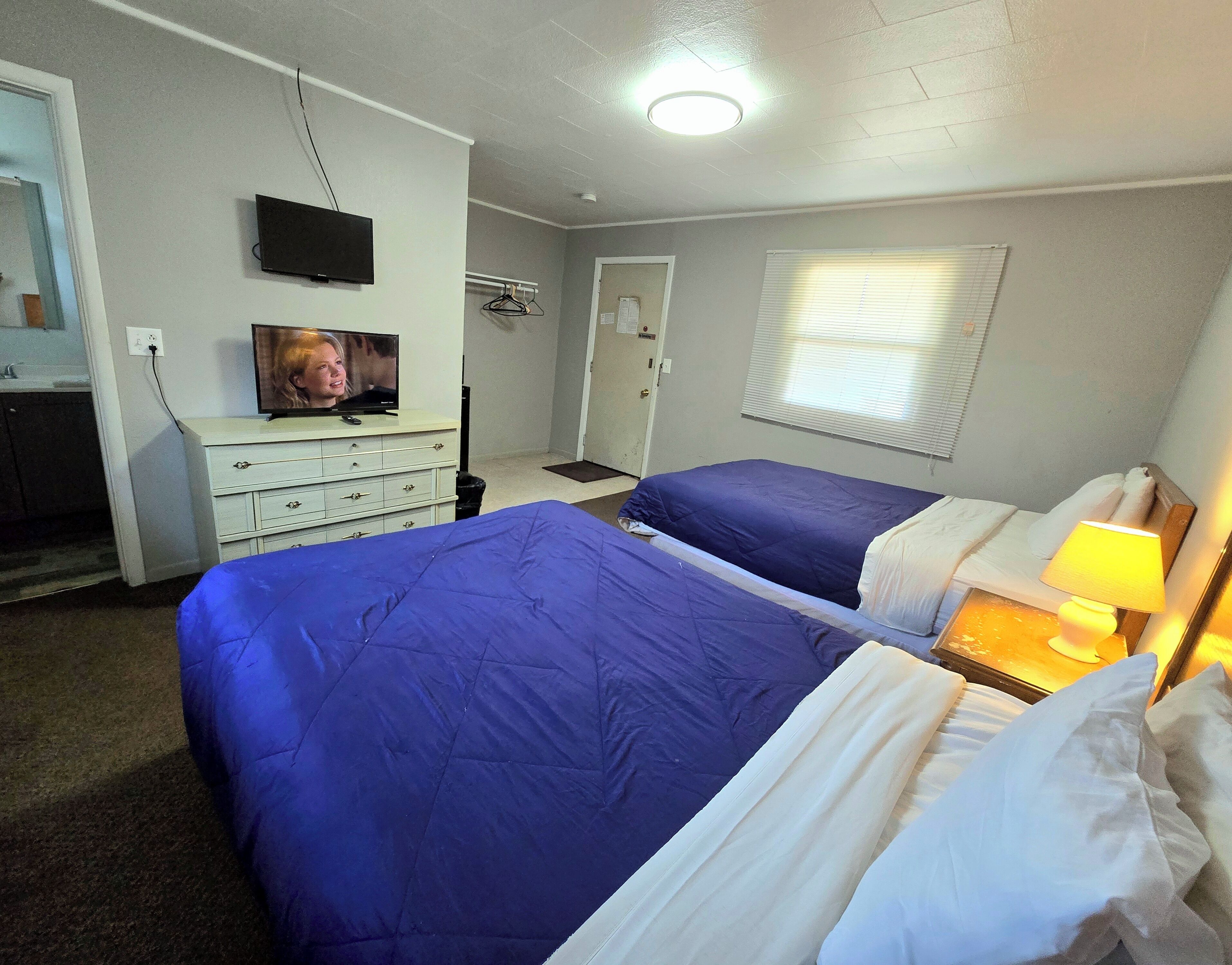 A hotel room with two beds, a television, a dresser, and a window with blinds.
