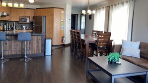 Una cocina moderna con barra, armarios de madera y electrodomésticos de acero inoxidable. Cuenta con un comedor con mesa y sillas, y una sala de estar con sofá y mesa de centro.