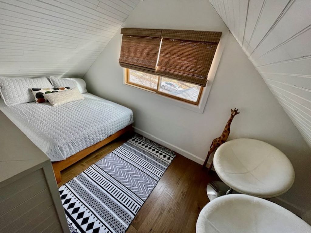 Een compacte slaapkamer met een bed, een raam met zonwering, een witte wastafel en een vloerkleed met dessin.