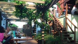 Et udendørs siddeområde med træborde og bænke, omgivet af grøn vegetation og en træpergola.