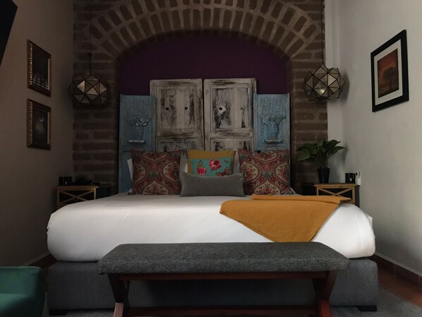A bedroom with a large bed, two nightstands, a bench, and a brick wall arch. - Hotel Casamada (San Miguel de Allende)