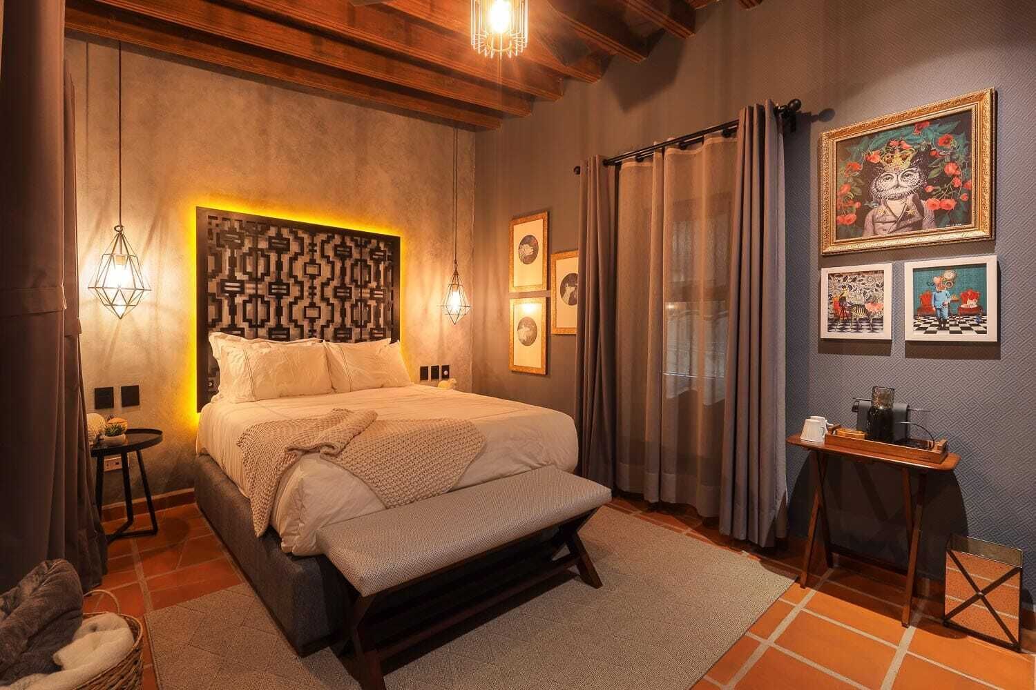 Een slaapkamer met een groot bed, een hoofdbord met een geometrisch patroon, twee wandlampen, een klein tafeltje met een lamp, en kunstwerken aan de wanden.