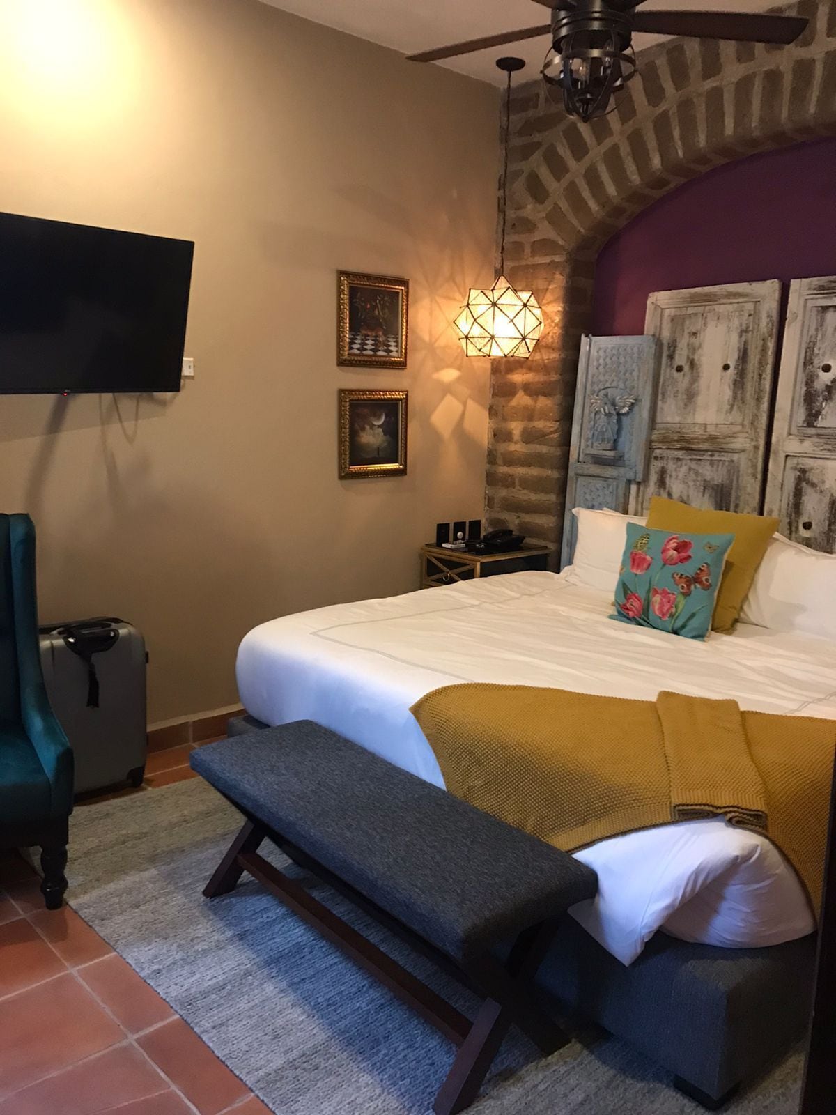 A hotel room with a bed, bench, suitcase, and a television on the wall.