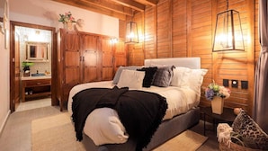 Une chambre confortable avec un plafond en bois, un grand lit agrémenté d’oreillers à motifs, une table de chevet avec un vase de fleurs et une armoire en bois.