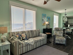 A living room with a striped sofa, a chair, a small table, and a lamp. There is a window with blinds, a ceiling fan, and artwork on the wall.
