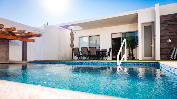 Ein moderner Außenpool mit einer Pergola, einem Essbereich mit Stühlen und einer Tür zu einer überdachten Terrasse.