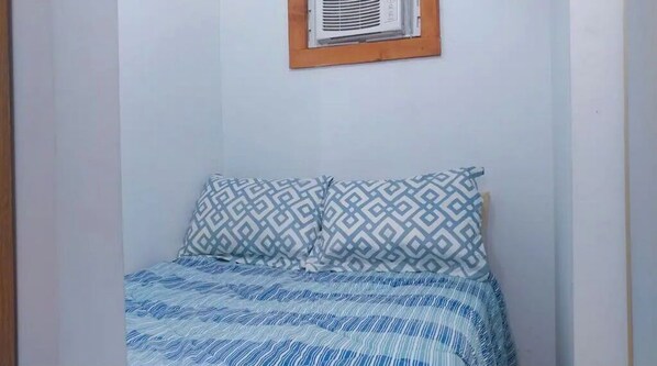 Una habitación pequeña y sencilla con una cama, dos almohadas estampadas y un ventanal con marco de madera.