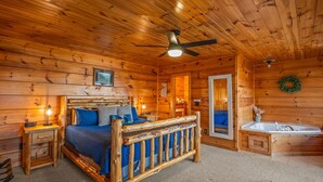 Een slaapkamer in een blokhut met een houten bed, een plafondventilator, een badkamer met wastafel en badkuip, en een open haard.